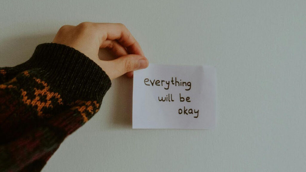 A hand in a patterned sweater holds a note with the handwritten message "everything will be okay" against a plain wall, conveying reassurance and hope.