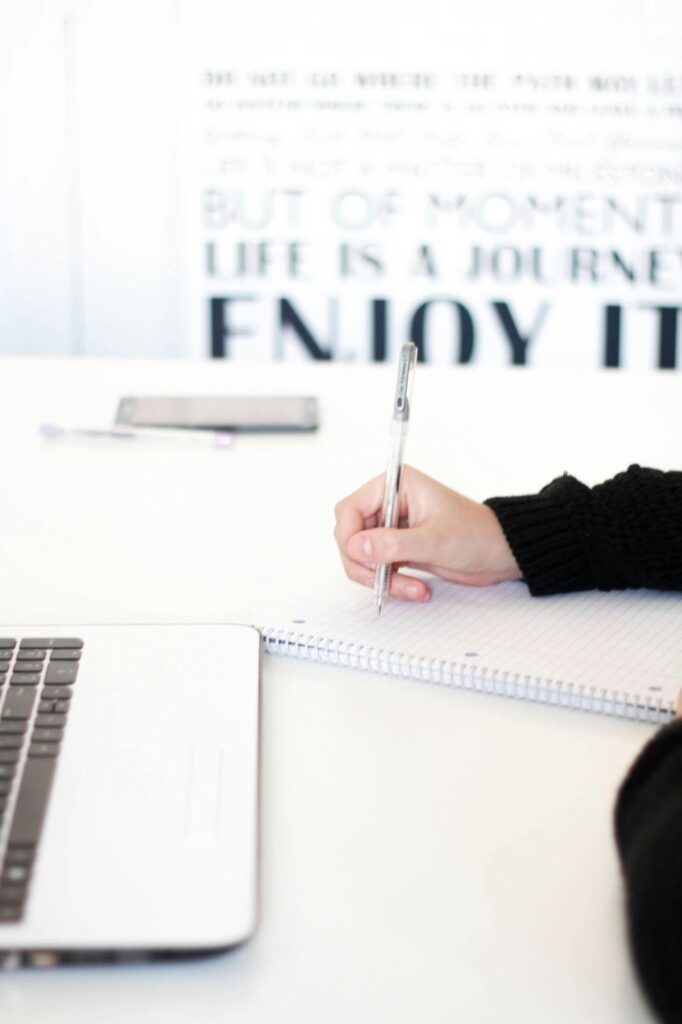 Person Writing With Pen and Laptop