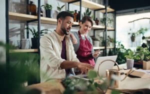 Two business owners in their flower shop looking at a laptop on a table and smiling.