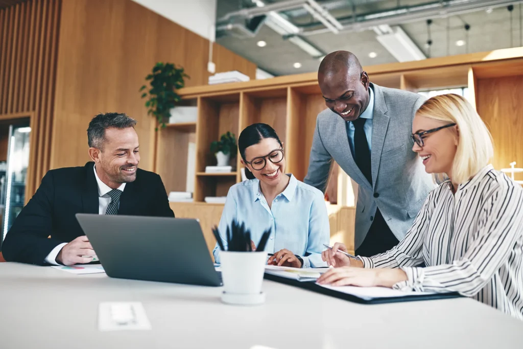 A group of office colleagues sitting around a table, smiling and looking at a laptop.